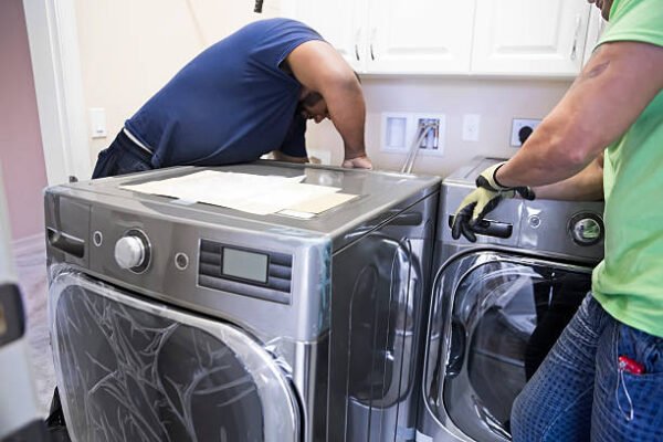 ll Two appliance delivery men are installing a new washer and dryer in the laundry room of home. One of the repairman is making connections in the back of the machine while his helper watches him. The new washer and dryer are energy efficient. Shot taken with Canon 5D Mark 3.